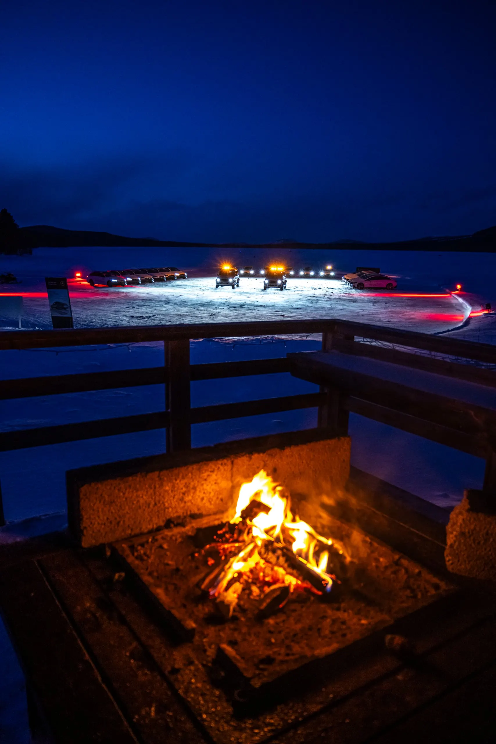 Feuerstelle am Seeufer, beleuchtete Fahrzeuge auf dem Eis